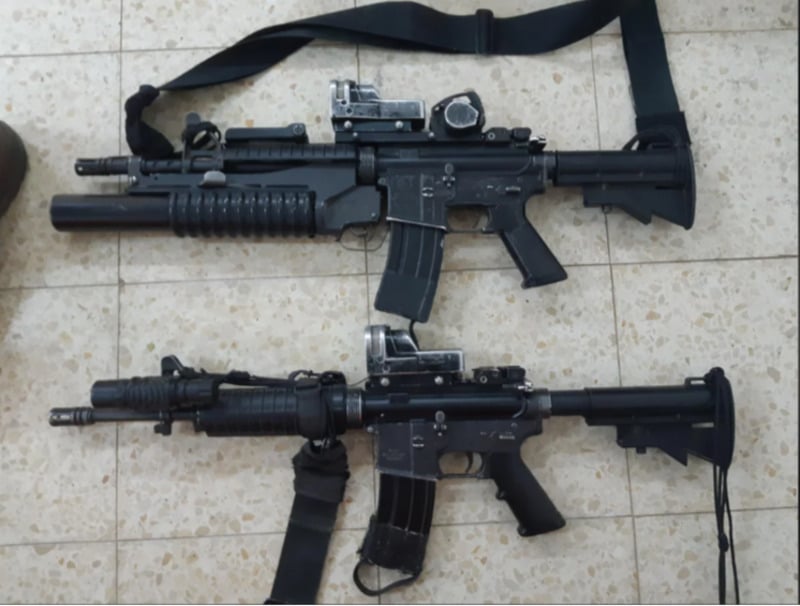 Two black assault rifles with scopes are laid side by side on a tiled floor. The top rifle has an attached grenade launcher and a sling, while the bottom rifle features a shorter barrel and also has a sling.