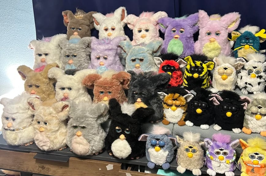 A collection of over twenty Furbies in various colors and styles is displayed on a table in several rows, with a dark blue background curtain behind them.