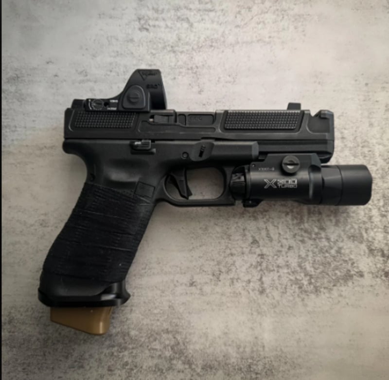 A black semi-automatic handgun with a textured grip, mounted flashlight, and a red dot sight, placed on a light, textured surface.