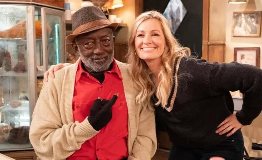 An older man in a hat and red shirt sits and points while smiling, next to a smiling woman with long blonde hair who has her arm around him, inside a cozy room with a refrigerator in the background.