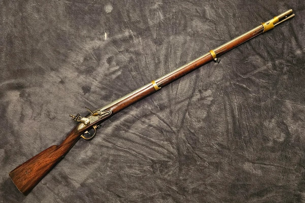 An antique flintlock musket with a wooden stock, metal barrel, and brass fittings is displayed on a dark textured surface.