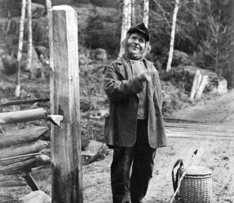 An older woman wearing a coat, scarf, and cap stands on a dirt road near a wooden fence and basket, with birch trees in the background. The photo is in black and white.