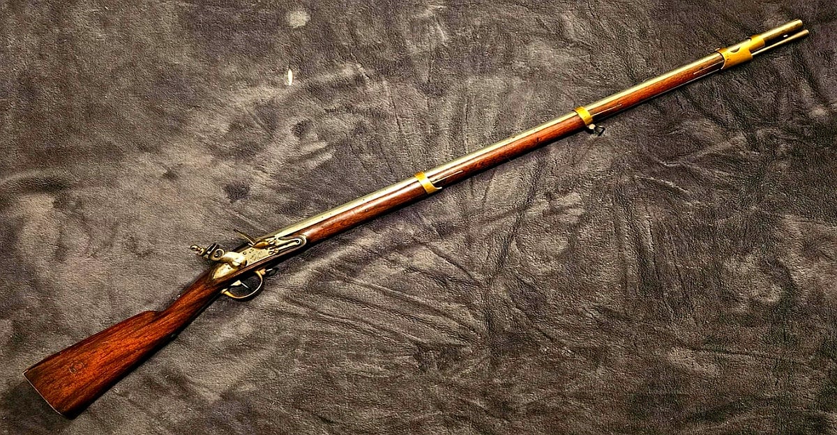 A vintage flintlock musket with a wooden stock, metal barrel, and brass fittings, displayed diagonally on a textured dark surface.