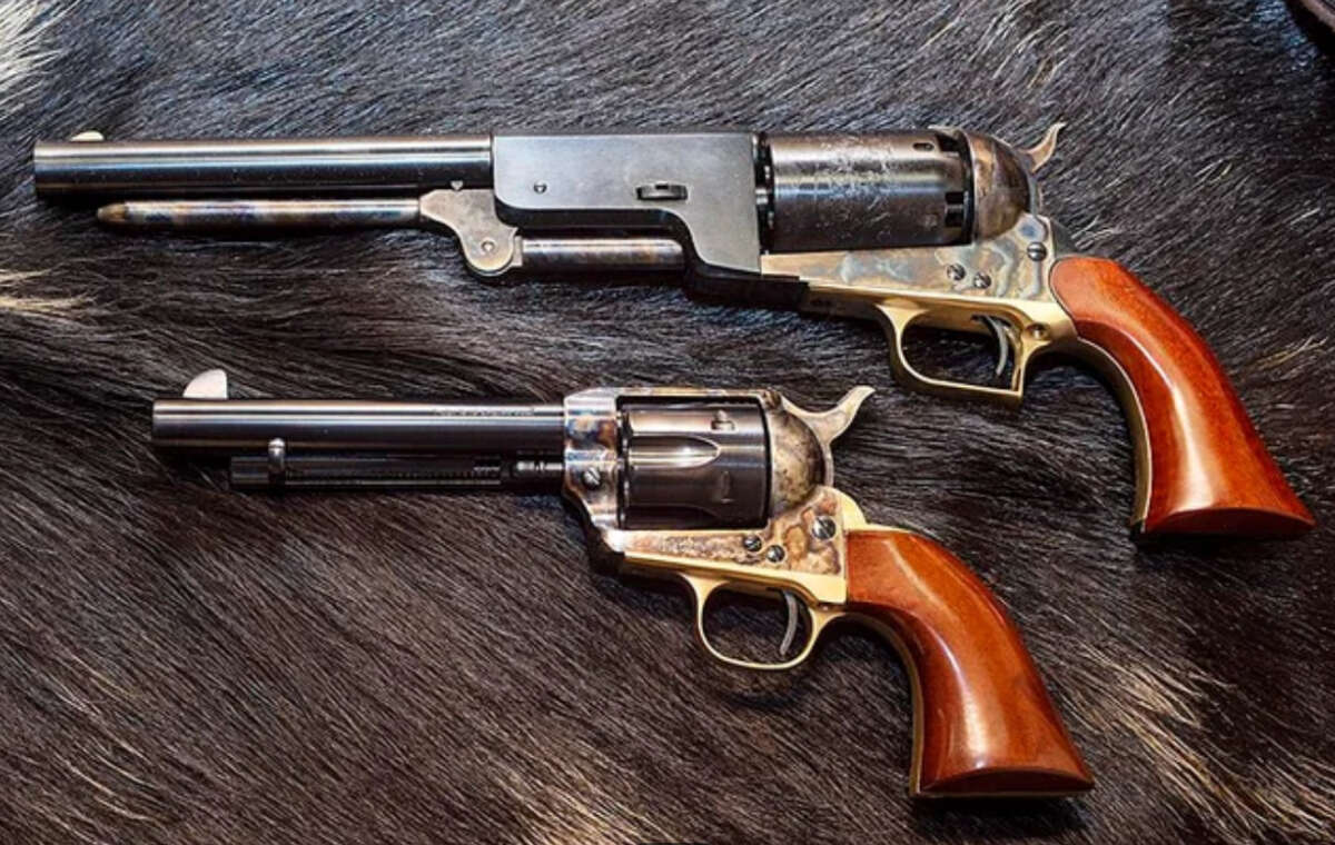 Two vintage revolvers with wooden grips and metallic finishes are displayed side by side on a dark, textured fur surface. The guns differ in length and design details.
