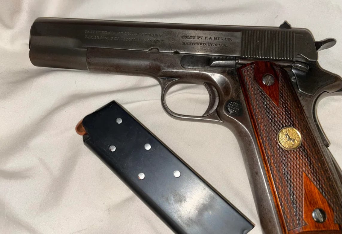 A Colt 1911 semi-automatic pistol with a wooden grip is lying on a light-colored fabric. The magazine is removed and placed next to the handgun.