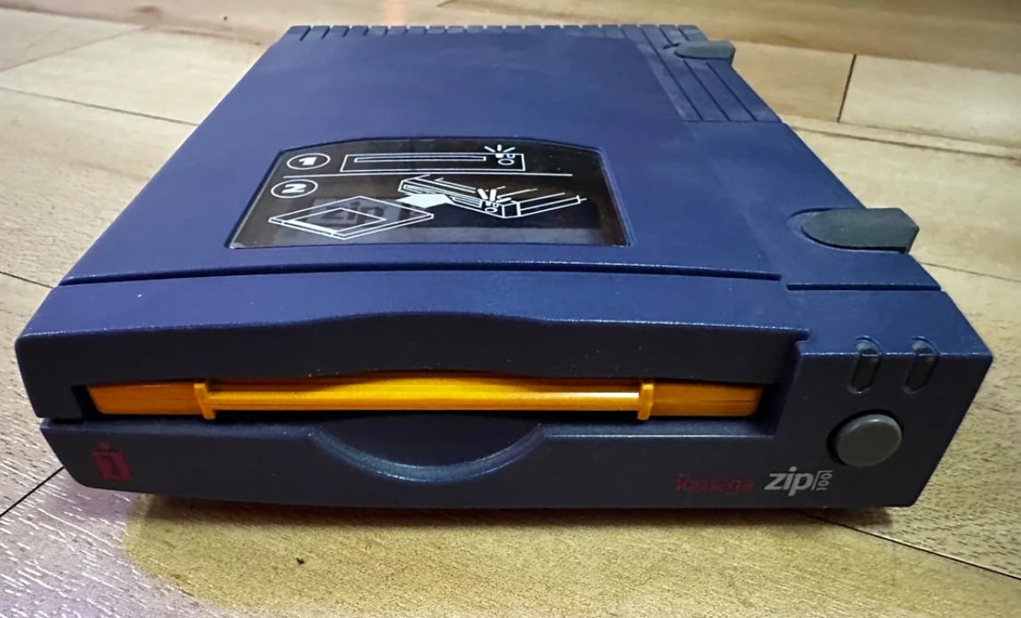 A close-up of a blue Iomega Zip disk drive on a wooden surface, with an illustrated label on top showing disk insertion instructions. The drive’s front slot is visible with a yellow tab inside.
