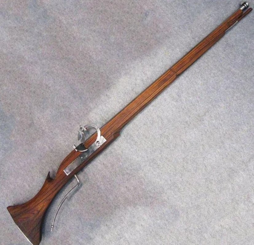 A matchlock musket with a wooden stock, metal trigger mechanism, and a long barrel, displayed on a neutral gray background.