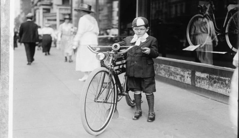 A young boy in vintage clothing and a cap stands on a city sidewalk holding a bicycle. He looks serious, and people walk by in the background near a shop window displaying another bicycle.