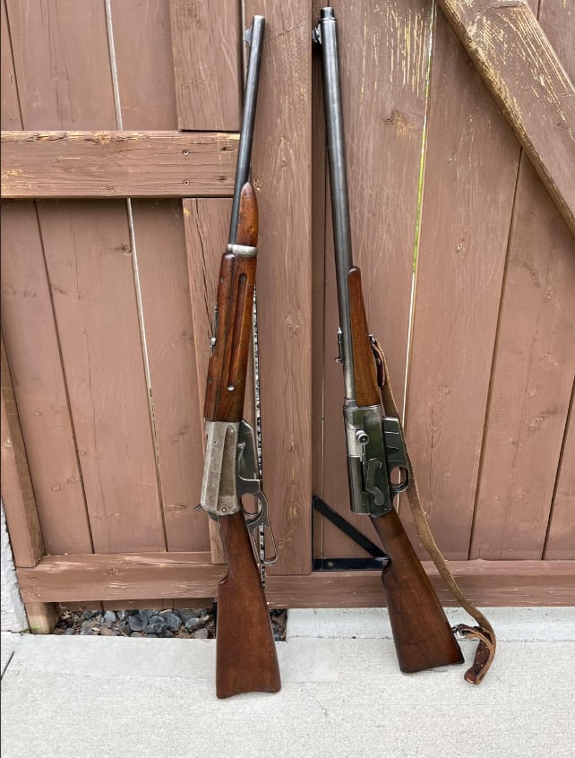 Two vintage rifles with wooden stocks leaning against a brown wooden fence outdoors, with a concrete surface beneath them. One rifle has a strap attached.