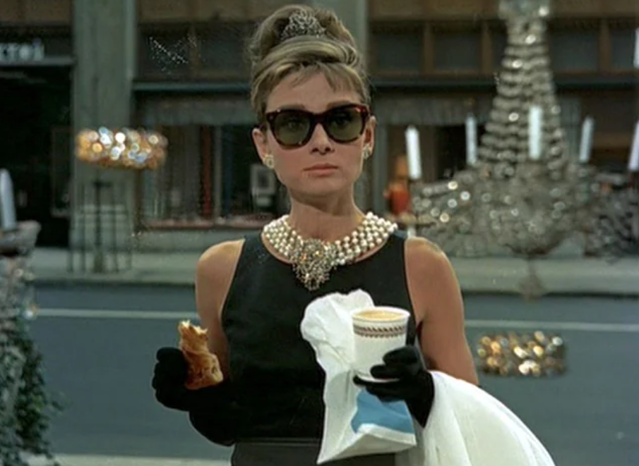 A woman wearing a black dress, pearl necklace, tiara, and large sunglasses stands on a city sidewalk holding a pastry and a coffee, with a jewelry store window display behind her.