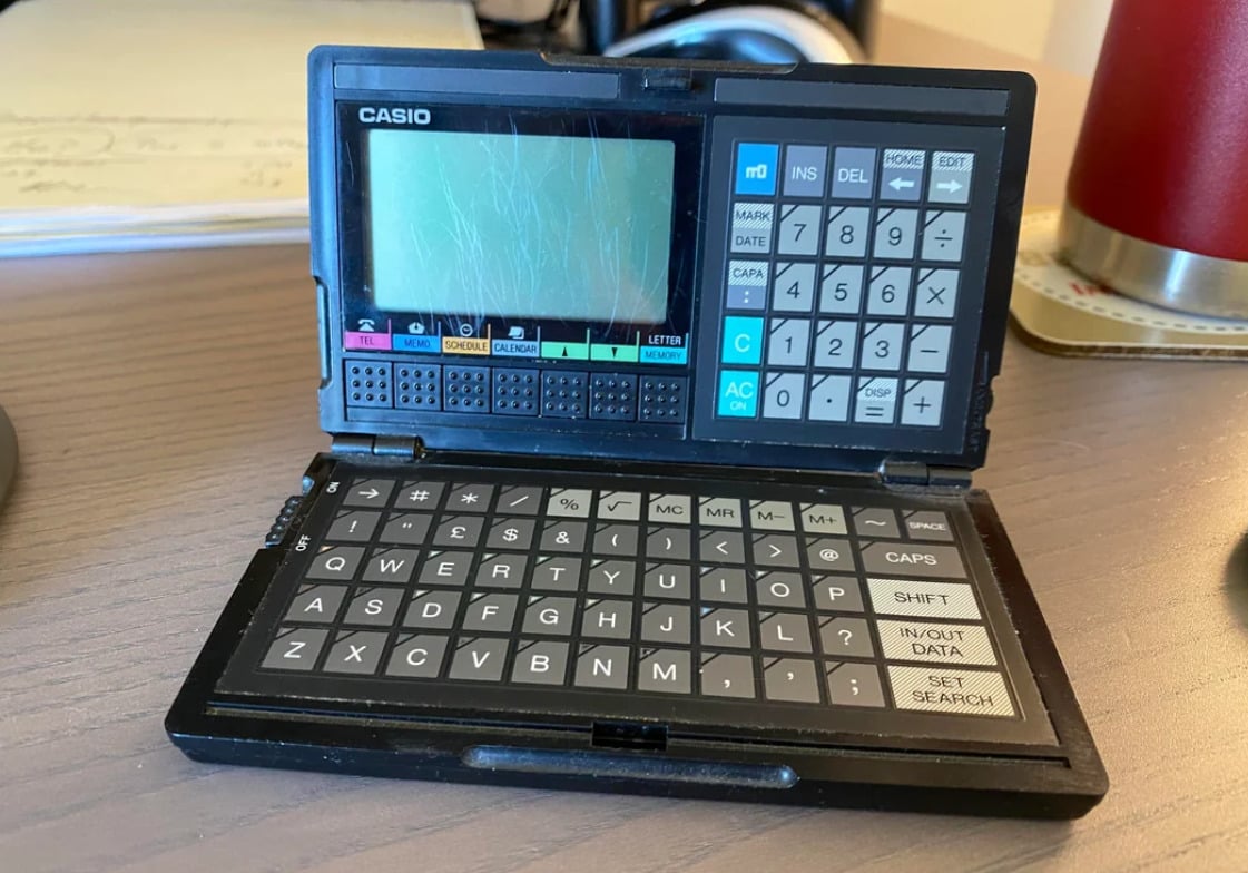 A vintage Casio electronic dictionary with a scratched screen sits open on a wooden desk, showing a keyboard and function buttons. Papers and a red cup are partially visible in the background.