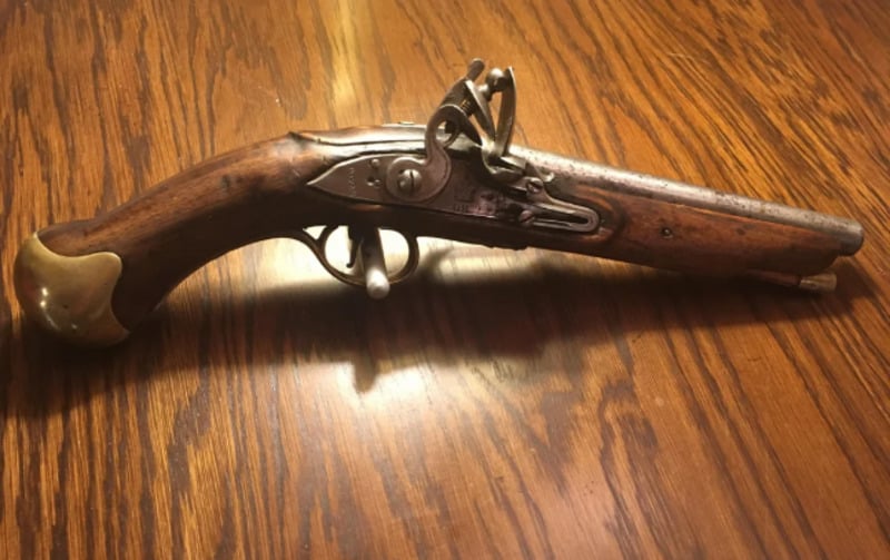 An antique flintlock pistol with a wooden handle and brass accents rests on a polished wooden table.