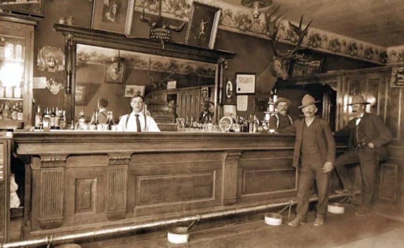 Sepia-toned photo of an old-fashioned bar with a long wooden counter, bartender behind it, and three men in hats and suits standing in front. Animal heads and framed pictures decorate the walls.