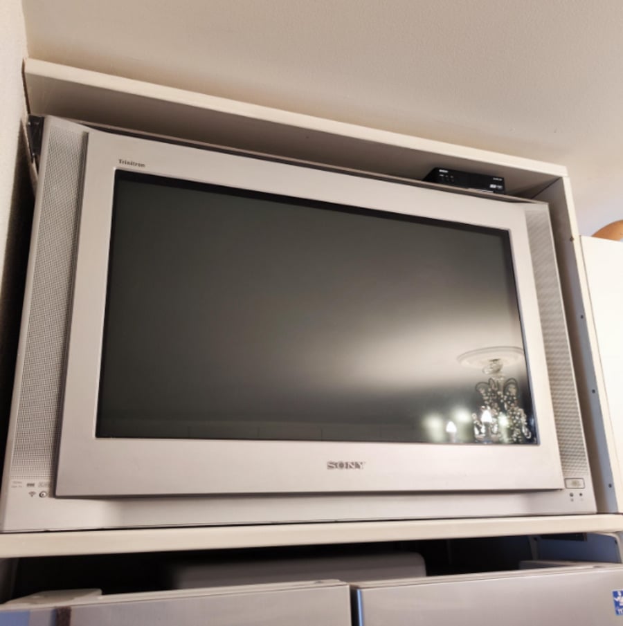 A silver Sony CRT television is placed on a shelf, with its screen turned off. The reflection of a light fixture can be seen on the display.
