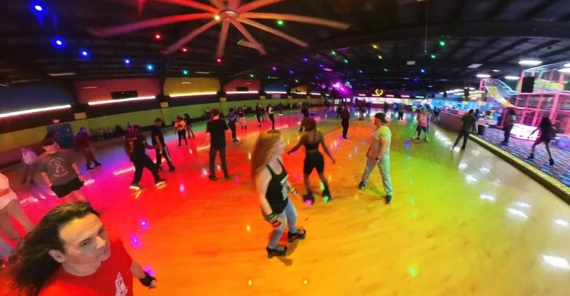 People roller skating on a brightly lit indoor rink with colorful lights reflecting on the floor. The rink is busy, and there are various skaters of different ages enjoying themselves. A large ceiling fan is visible above.