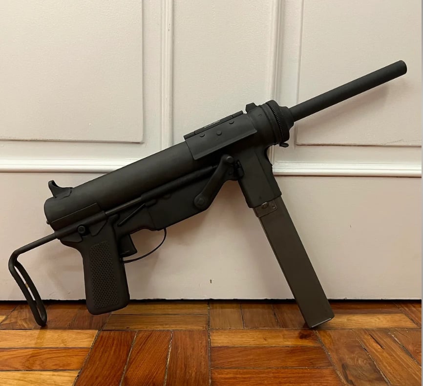 A black submachine gun with a folding stock and long magazine is propped against a white paneled door on a wooden floor.
