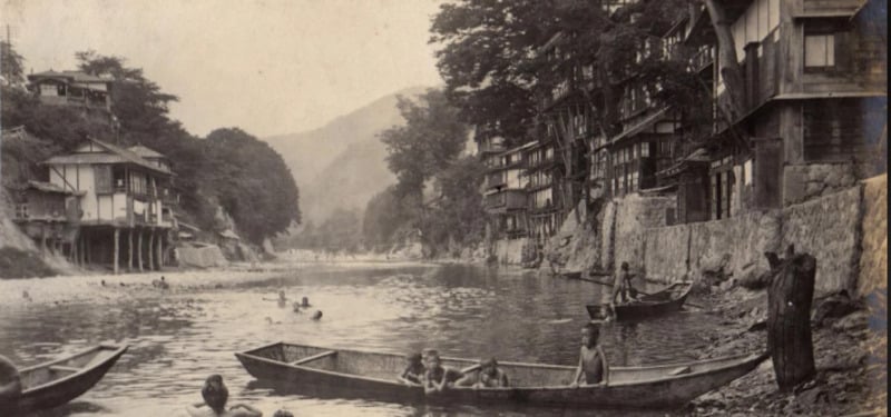 Children swim and play in a river alongside wooden boats, with stilt houses lining the riverbank and trees in the background. The image is in black and white, evoking a historic or early 20th-century setting.