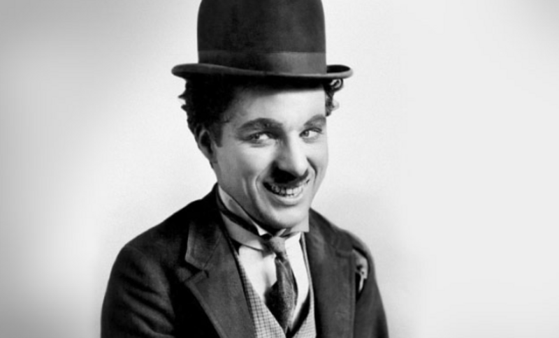 A black and white photo of a man with a small mustache, wearing a bowler hat, a suit, and a bowtie, smiling at the camera against a plain background.
