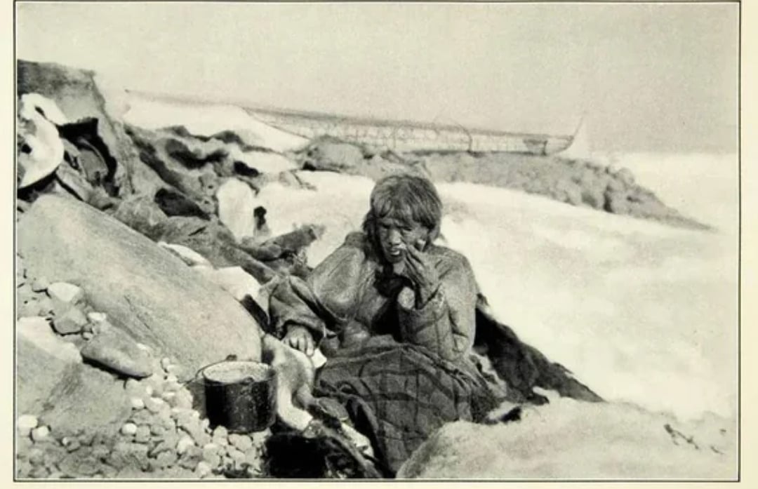 A woman sits on rocky ground, wrapped in a blanket, appearing distressed. Snow and ice surround her, with a pot nearby and a ship stranded in the background amid a harsh, frozen landscape.