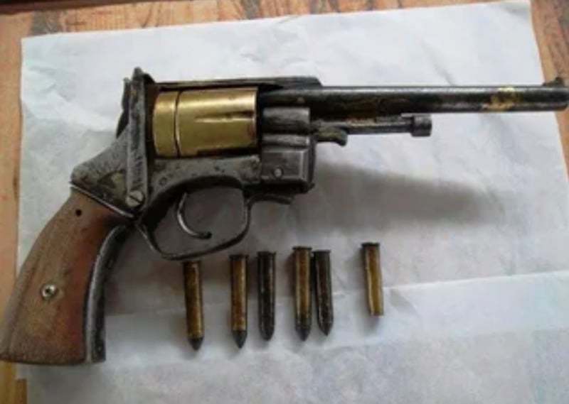 A revolver with a wooden grip and brass cylinder is placed on a white surface. Five bullets are arranged in a row beneath the gun.