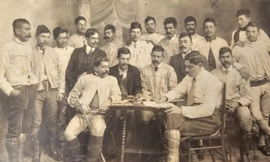 A sepia-toned vintage photo showing a group of men, some in uniforms and others in suits, gathered around a table. Two men sit and write while others stand behind them, posing for the camera in a formal setting.