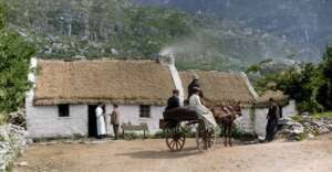 A colorized photo of people in early 20th-century clothing gathered outside white thatched cottages; some stand near a table, while others are seated on a horse-drawn cart, with mountains in the background.