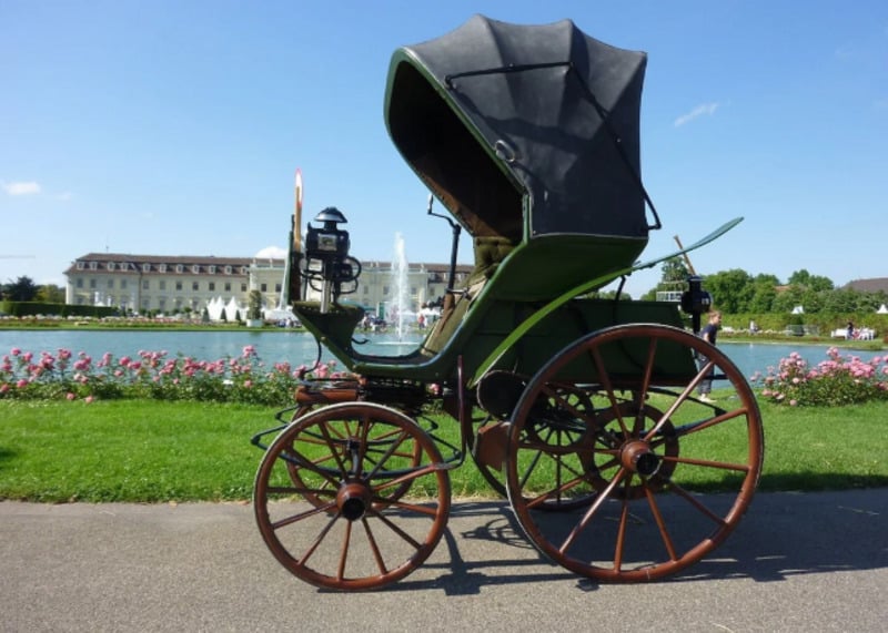 A vintage green horse-drawn carriage with large wooden wheels is parked on a path by a garden and pond, with a grand white building and fountains in the background under a clear blue sky.