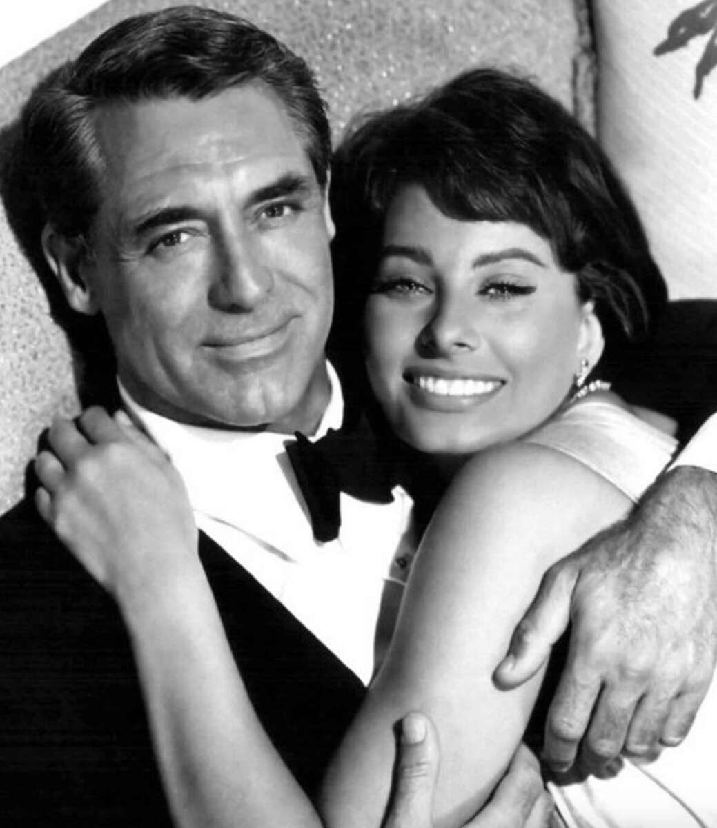 A man in a tuxedo and a woman in a sleeveless dress smile and embrace as they pose closely together on a couch in this black-and-white photo.