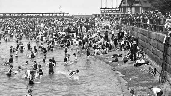 A crowded beach scene from the early 1900s shows people swimming, wading, and relaxing near a pier and a large building, with many wearing vintage swimsuits and hats.