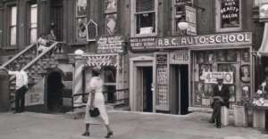 Black-and-white photo of a city street with people walking, a man sitting on a crate, and storefronts for a beauty salon, barber, and auto school. Posters and signs fill the building facades.