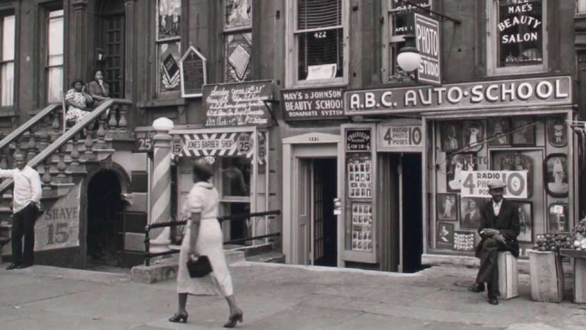 Black-and-white photo of a city street with people walking, a man sitting on a crate, and storefronts for a beauty salon, barber, and auto school. Posters and signs fill the building facades.