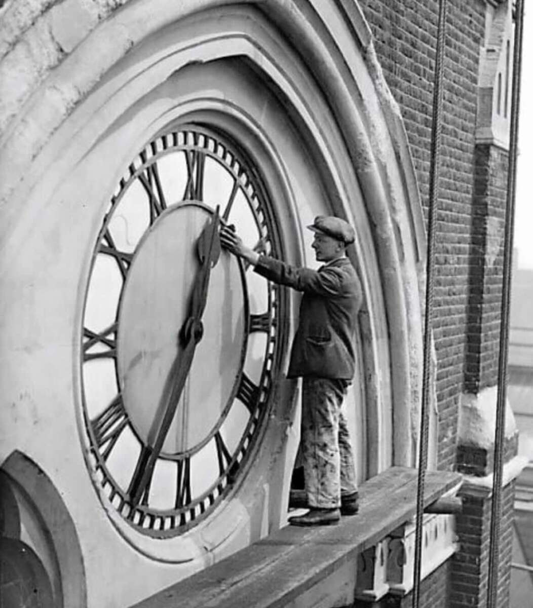 A man in a cap and jacket stands on a narrow ledge, adjusting the hands of a large clock on the exterior of a brick building.
