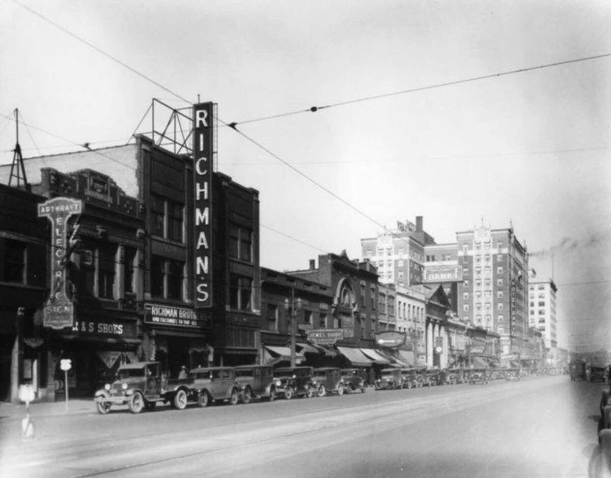 A black-and-white photo of a busy city street from the early 1900s, lined with vintage cars and storefronts. A tall sign reads "RICHMAN'S" and several buildings and shops are visible along the road.
