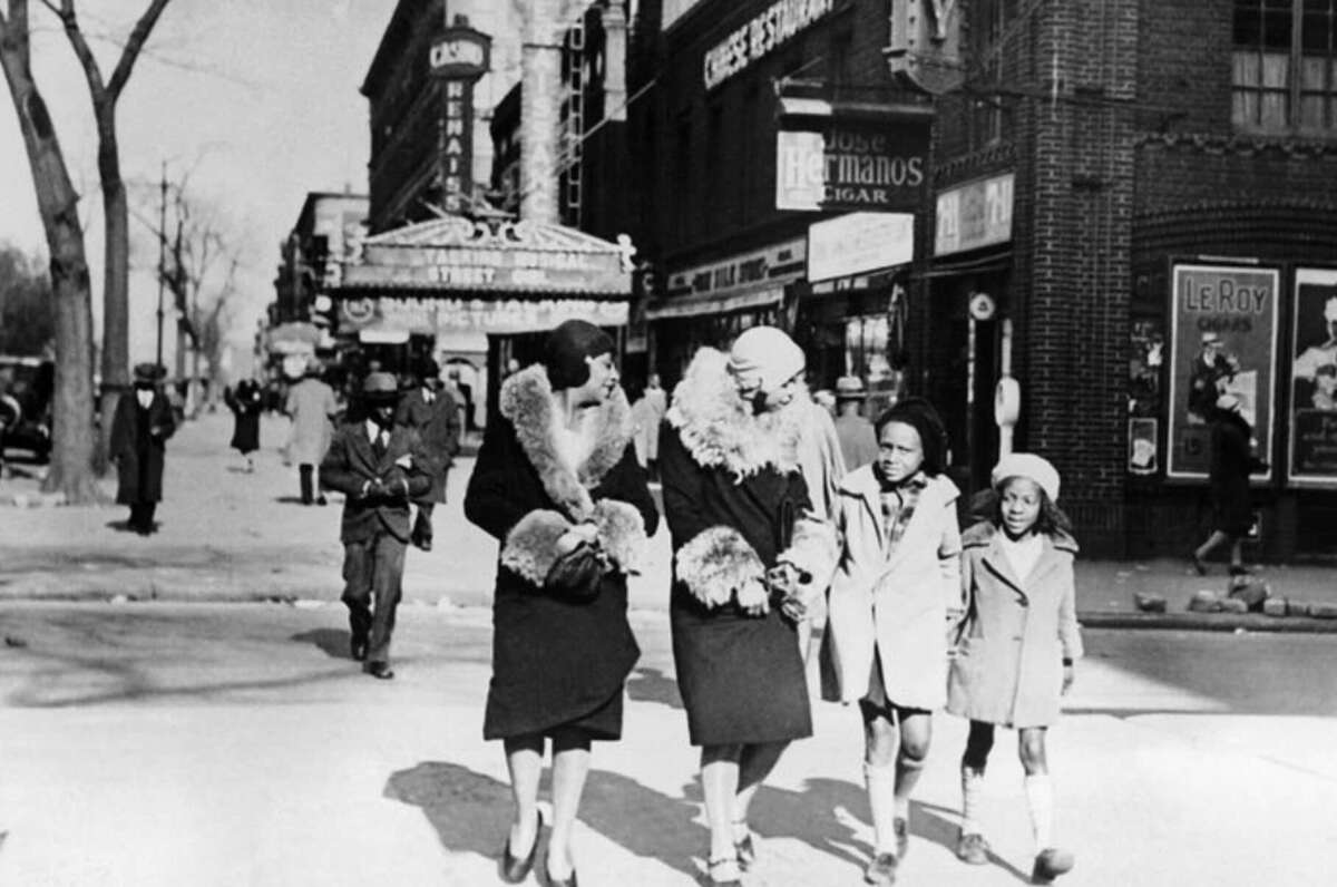 Two women in fur-collared coats walk arm in arm with two children on a city street, lined with shops and a theater marquee, in a black-and-white photo from the early 20th century.