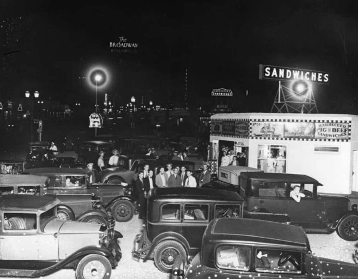 A black-and-white photo shows a busy 1930s drive-in diner at night, with vintage cars parked and people gathered near a brightly lit sandwich stand under large neon signs.