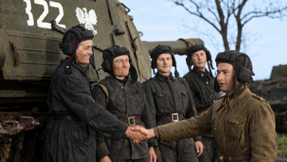 Five men in military uniforms stand near a green tank; two in the foreground shake hands while three others smile behind them. The tank has the number “322” and a white eagle symbol painted on it.