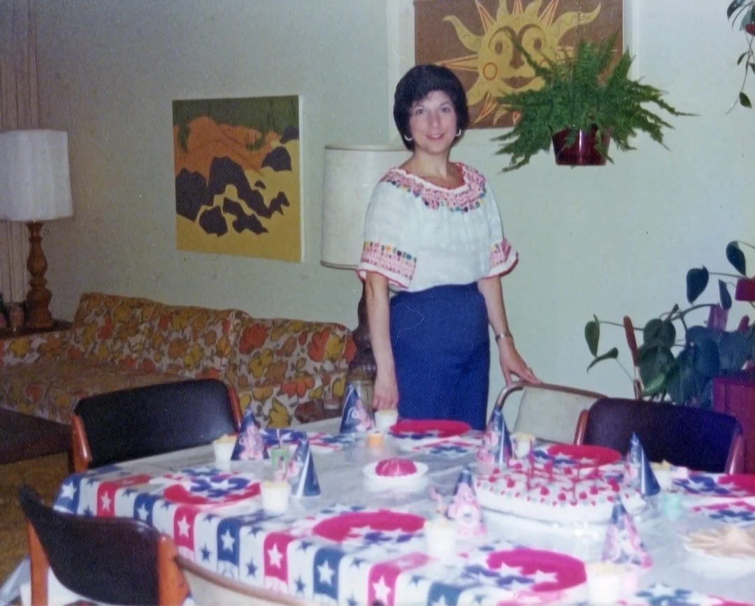 A woman stands by a festively decorated table set for a party, with patriotic-themed tablecloth, hats, and cake. The room has colorful wall art, a sofa with a floral pattern, and various houseplants.
