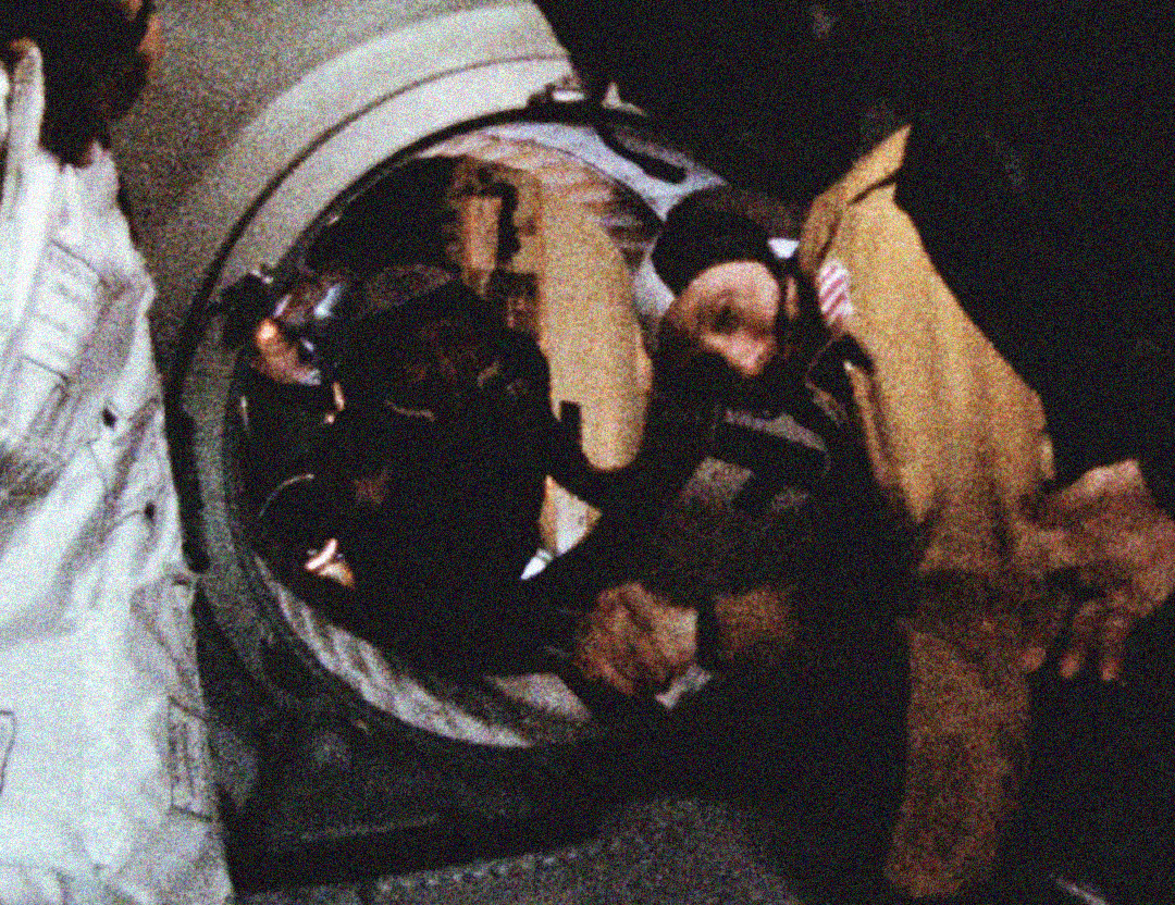 A grainy photo shows two astronauts, one in a brown suit and one in a white suit, shaking hands through a circular hatch between two spacecraft.