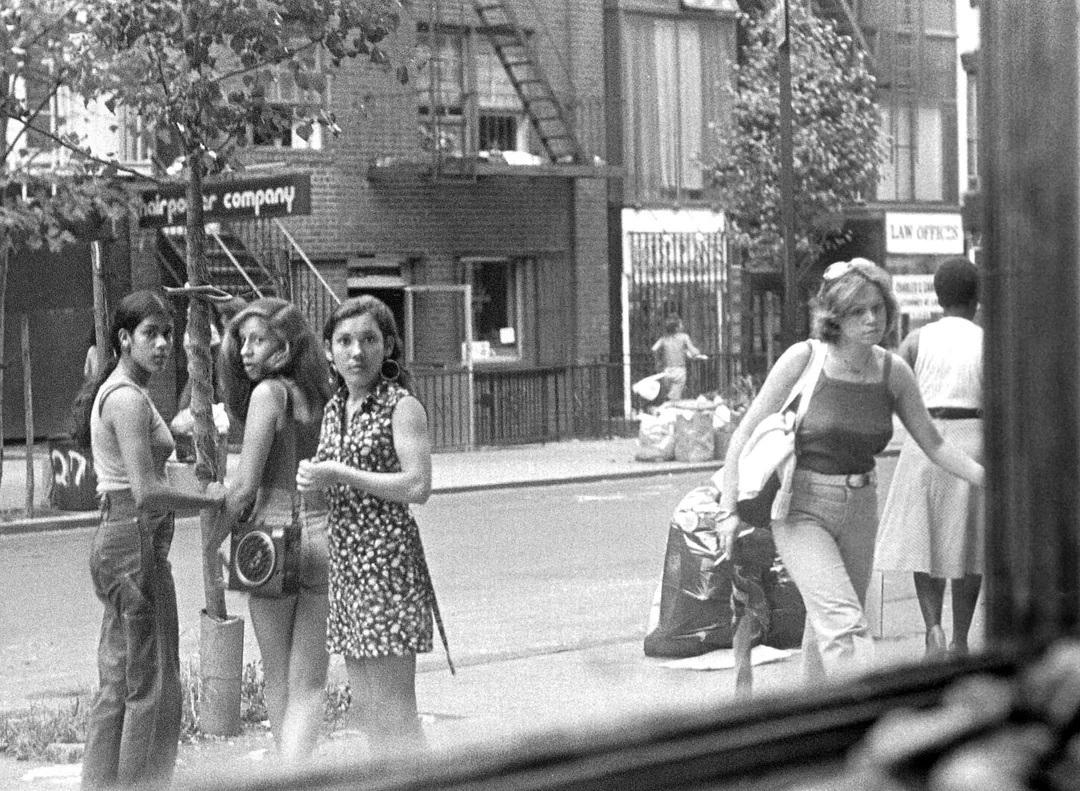 Black and white photo of four young women standing on a city sidewalk, looking toward the camera, with buildings, trees, and people in the background.