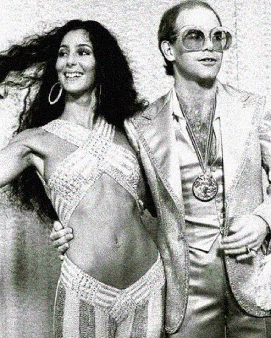 A woman in a sparkling cut-out outfit and hoop earrings stands next to a man wearing glasses, a shiny suit, and a large medallion; both are smiling and posing together.