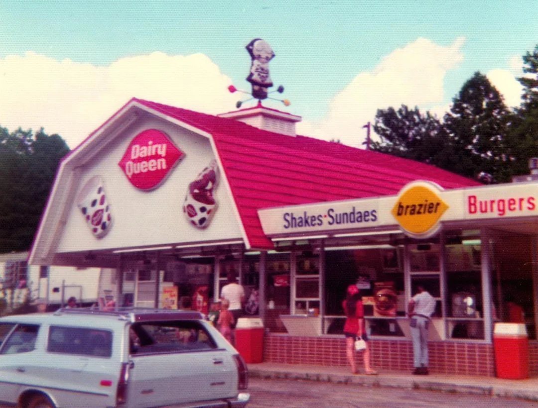 A vintage Dairy Queen restaurant with a red roof, large signs, and a cartoon figure on top. People stand at the order window, and a white station wagon is parked in front.