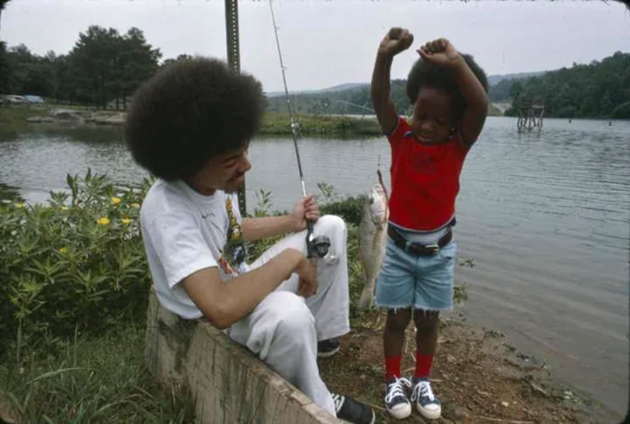 A man holding a fishing rod sits by a lake with a caught fish, while a smiling child in a red shirt and denim shorts stands nearby, cheering with arms raised in excitement. Trees and water are in the background.