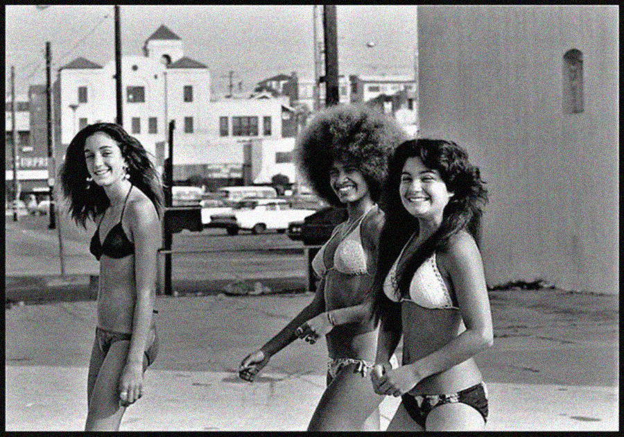 Three young women in bikinis walk together, smiling, on a sunny urban street with vintage cars and buildings in the background. The photo is in black and white, evoking a retro or 1970s vibe.