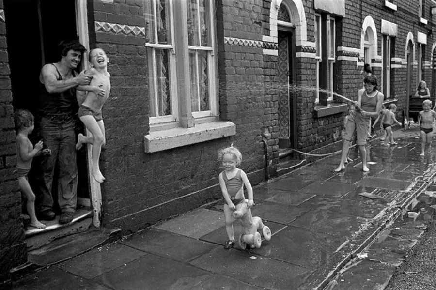 Children in swimsuits laugh and play with water on a wet sidewalk outside brick row houses, while an adult helps a child out a doorway. Another child rides a toy horse, and others spray water from hoses.