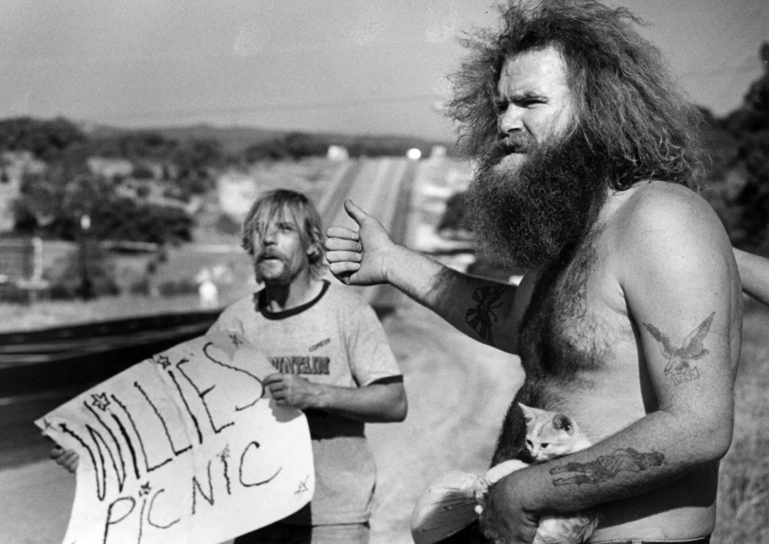 Two men stand by a rural road; one shirtless with a bushy beard and tattoos holds a kitten and thumbs for a ride, while the other holds a handmade “Willie’s Picnic” sign. The background shows an empty highway.