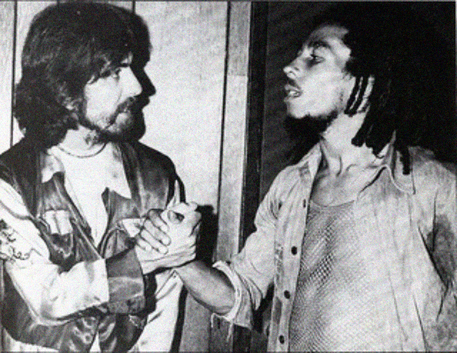 Two men greet each other with a handshake, looking at each other. One has long hair and a beard, wearing a jacket; the other has dreadlocks and a mesh shirt. The image is black and white and has a candid, friendly feel.