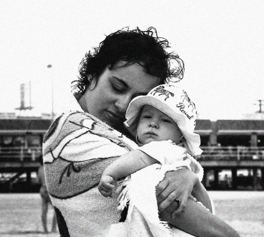 A woman holds a baby wrapped in a towel at the beach. The baby wears a sun hat and looks at the camera, while the woman rests her head gently against the baby, with a pier visible in the background.