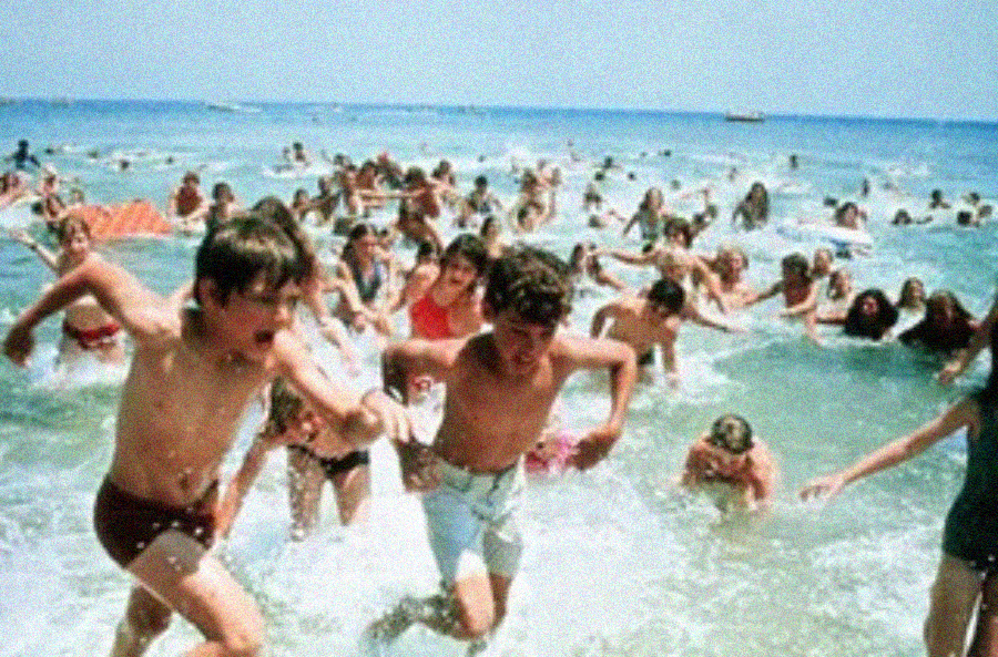 A large crowd of people in swimsuits rushes out of the ocean in apparent panic, splashing through shallow water under a clear blue sky.
