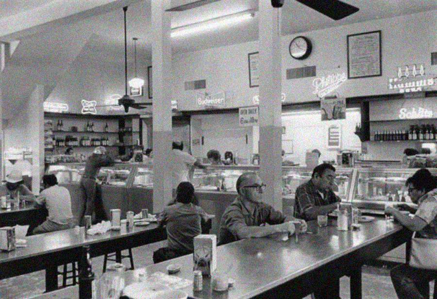 Black and white photo of a busy diner with people sitting at long tables eating and drinking; several staff serve food behind a counter lined with neon signs and shelves.