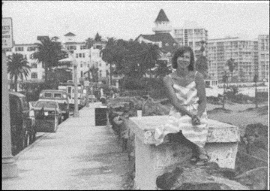 A woman in a striped dress sits on a stone railing alongside a sidewalk, with cars parked on the street and buildings, including a distinctive turreted structure, in the background. The image is black and white.